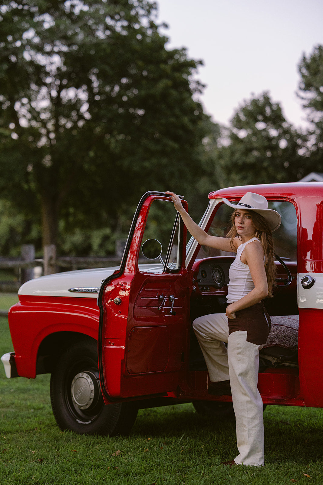 Woman in a cowboy hat standing next to a red truck in a grassy area with trees in the background